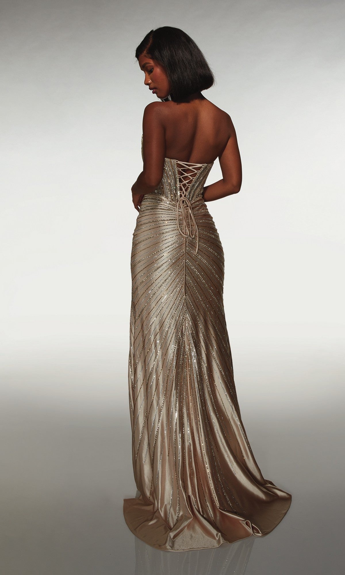 A woman faces away, wearing the Alyce 62142 long prom dress: a strapless metallic gold satin gown featuring a lace-up corset bodice and shimmering textured pattern.